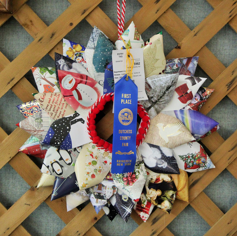 A photo of an award-winning arts & crafts project from the Dutchess County Fair, a wreath made from festive wrapping paper. The wreath has a "First Place" blue ribbon on it from the Dutchess County Fair.