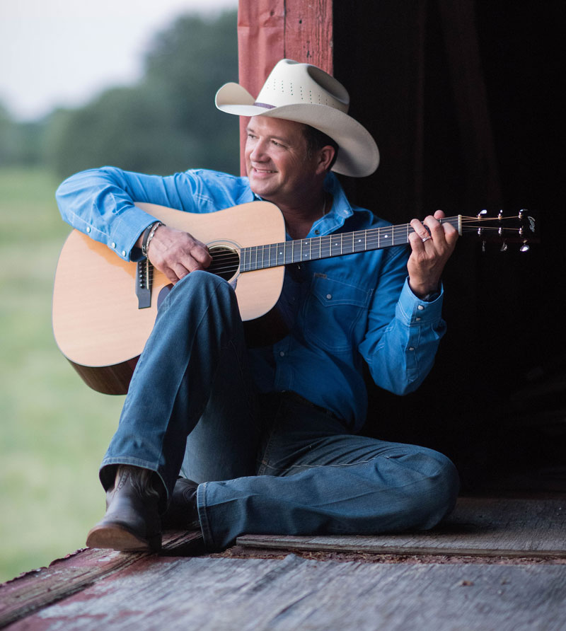 A photo of the musician Tracy Byrd seated in rustic surrounding playing an acoustic guitar. Tracy Byrd is a performer at the 2024 Dutchess County Fair.